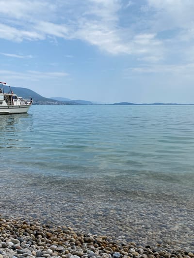 The image shows a serene waterscape scene with a clear sky and calm water. In the foreground, a pebbled shore is visible, with the water gently lapping at the stones. The water is clear enough to see the pebbles beneath the surface. A white boat with a red canopy is docked on the left side of the image, its reflection visible in the water. In the distance, rolling hills with scattered buildings form the backdrop, suggesting a coastal town or settlement. The overall atmosphere is tranquil and picturesque, evoking a sense of peace and natural beauty. The scene is likely a waterfront location in Cortaillod, Switzerland, known for its scenic views of Lake Neuchâtel.