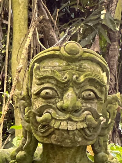 The image is a close-up, eye-level shot of a moss-covered stone statue of a mythical creature's head. The statue features exaggerated, stylized facial features including large, bulging eyes, a prominent nose, and a wide, open mouth displaying rows of sharp teeth. Ornate, swirling carvings adorn the forehead and cheeks, and what appear to be horns or headdresses extend from the sides of the head. The statue is positioned outdoors, with a background of dense foliage and tree trunks, suggesting a natural or garden setting. The overall color palette is dominated by greens from the moss and the surrounding vegetation, with brown tones from the stone and trees. No people or animals are visible, and no specific location in Ban Bang Khao, Thailand, can be determined from the image itself.