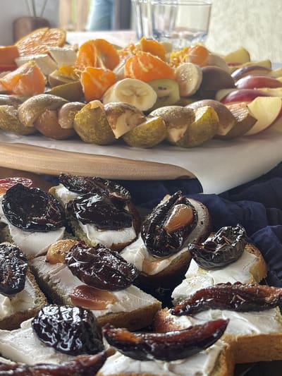 A close-up shot presents a wooden serving tray, likely located in Lahojsk selsaviet, Belarus, containing an assortment of fruit and canapés. On the left side of the tray, arranged in rows, are slices of apple, pear, kiwi, banana, and tangerine. A white, possibly wax paper, divider separates the fruit from the canapés on the right. The canapés consist of small, square pieces of bread topped with a white spread, possibly cream cheese, and dark-colored, shiny fruit, which might be prunes or plums. A dark blue, crumpled cloth is visible under the canapés, providing contrast to the food. The lighting suggests an indoor environment, and the depth of field is shallow, focusing primarily on the food items. No people are directly visible in the frame.
