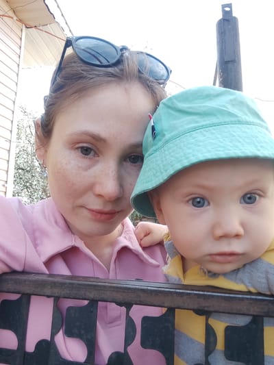 The image is a selfie featuring an adult female and a baby. The woman has fair skin, freckles, and her hair is pulled back with sunglasses perched on her head. She is wearing a pink collared shirt. The baby, positioned to her right, is wearing a light blue hat and a yellow and gray striped shirt. They are both leaning against a dark metal fence. Behind them, the background shows a building with light-colored siding and what appears to be a blooming tree. A dark pole is also visible. The location is likely outdoors, potentially in the city of Malaya Purga, Russia, based on the prompt.