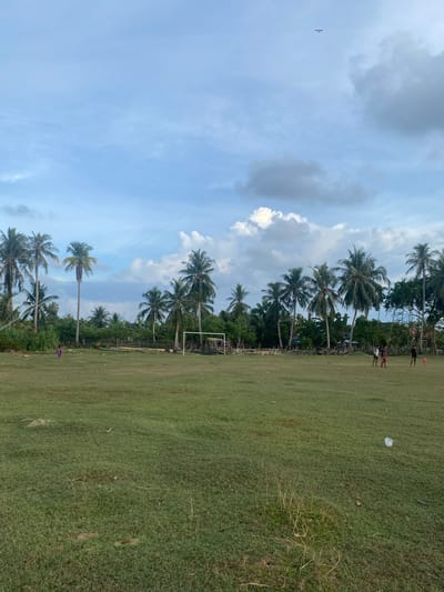 The image depicts an outdoor scene in Keudeublang, Indonesia, under a partly cloudy sky. In the foreground, a large, grassy field occupies the majority of the frame, with some patches of dry grass and scattered debris, including a white plastic bag. Several palm trees are visible in the background, forming a dense line along the edge of the field. Further back, there appears to be a fence or barrier made of wood and fencing material, and a rudimentary soccer goal with white posts. A few people are scattered across the field; on the left, a small figure in a purple garment is walking, while on the right, three darker-clothed figures, possibly young men, are standing near the goal. One of the figures on the right appears to be kicking a ball. A very small figure, possibly a child, is also visible to the far right. The sky above is a mixture of blue and gray clouds, with a thin, dark line stretching diagonally across it from the upper left to the middle, possibly indicating a kite string.