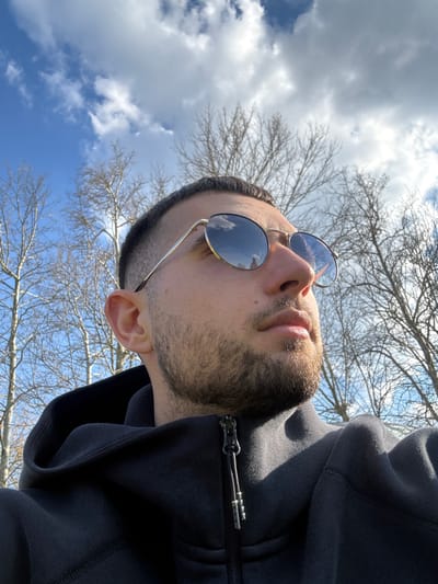 A man is pictured from a low angle, looking upwards. He is wearing round sunglasses with gold frames. He has short, dark hair and a beard. He is wearing a dark hoodie, partially zipped up. Above him, there is a clear blue sky with white clouds, and bare tree branches. The photograph may have been taken outdoors on a bright day. The location is not evident from the image and cannot be pinpointed to Dushevo, Bulgaria.