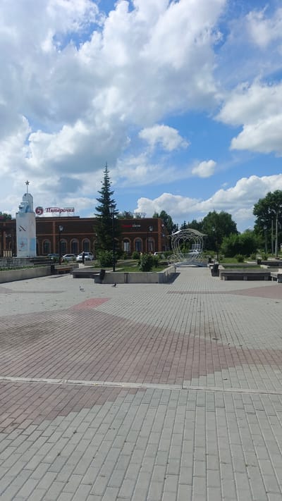 An exterior daytime, vertical, eye-level shot shows a public square in Kuybyshev, Russia. The square is paved with red and grey interlocking bricks. A large building with a dark brick facade stands in the background, featuring arched windows and a prominent red sign with white Cyrillic lettering that reads "Пятёрочка" (Pyaterochka), a Russian supermarket chain. To the left of the building is a white, monument-like structure with blue accents and a star at the top. In the center of the square, a large, white, ornate metal structure resembling a carriage or gazebo with decorative scrollwork stands on a paved walkway. Several trees, including a tall, slender evergreen tree, are planted in raised flower beds. Benches are visible in the background near the building and around the metal structure. The sky is blue with scattered white clouds. Two pigeons are visible on the pavement in the foreground. There are no people clearly visible in the frame.