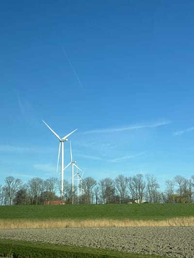 The scene is a rural landscape near Winkel, Netherlands, captured on a clear day under a predominantly blue sky. A distinct white aircraft contrail diagonally traverses the upper sky from the upper-left to the mid-right, with a subtle, wispy cloud formation visible lower in the mid-right sky.

In the mid-ground, a linear arrangement of at least four white, three-bladed wind turbines stands prominently. The leftmost turbine is the largest and closest, with its blades positioned in a stationary 'V' shape. The subsequent turbines diminish in size towards the right, suggesting distance. Behind these turbines, a dense horizontal line of leafless deciduous trees forms a barrier, obscuring the horizon. Partially visible behind the tree line are the upper sections of a red-tiled roof and other light-colored building structures.

The foreground is composed of three distinct horizontal bands: on the far left, a section of tilled, dark brown earth; adjacent to it, a strip of lighter brown, possibly dry vegetation or reeds; and finally, a broad, elevated green grassy embankment or dike stretching across the entire width of the lower image. No people or animals are visible.