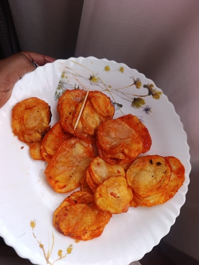 A close-up image depicts a white, scalloped-edge plate containing approximately 15-20 round, thinly sliced, fried food items, consistent with potato crisps or thinly fried potatoes. These items are coated in a reddish-orange spice or sauce, with some displaying small dark specks. A single light-colored wooden toothpick is partially inserted into one of the food items in the upper central portion of the plate. A dark-skinned human hand, adorned with a ring on the ring finger, is visible in the upper left, holding the rim of the plate. The background is an out-of-focus, light-colored, possibly mauve or light brown, textile surface, resembling a curtain or fabric wall covering. The specific location in Kahawa Wendani ward, Kenya, cannot be determined from the visual evidence.