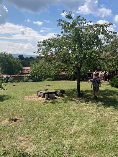 The image depicts an outdoor scene in the ethnographic complex of Zheravna, Bulgaria. The foreground features a grassy lawn with a stone table and several log seats arranged around it. A medium-sized tree with green leaves shades part of the lawn. In the mid-ground, a group of people, including adults and children, are walking away from the camera and towards buildings with traditional red-tiled roofs. The background shows more buildings, lush green trees, and a distant hilly landscape under a bright blue sky with scattered white clouds. The overall impression is a sunny day in a historic, rural setting.