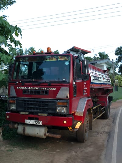 The image shows a red Lanka Ashok Leyland fuel tanker truck parked on the side of a road in Karandugoda, Sri Lanka. The truck is facing the viewer, with its front slightly angled to the left. The truck has a large, cylindrical tank mounted on its chassis, with the word "LUBRICANTS" visible on the side of the tank in white lettering. The truck's cabin is red, and it features the brand name "LANKA ASHOK LEYLAND" in white lettering above the grille. The license plate of the truck is visible and reads "WP LC - 7200".

The truck is parked on a dirt and gravel shoulder next to a paved road, which appears to be a two-lane road with a white line marking the edge. In the background, there are trees, palm trees, and a building with a tiled roof. The sky is overcast, suggesting it might be early morning or late afternoon. There are no people visible in the image. The overall scene depicts a stationary vehicle on a roadside in a rural or semi-rural setting.