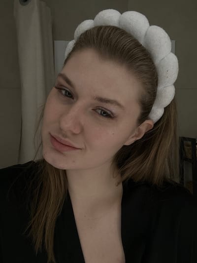 A young woman with fair skin and light brown hair pulled back in a ponytail poses at an angle. She wears a white, puffy headband. Her makeup includes a light shimmer on her eyelids and darkened lashes. She has a slight, subtle smile. She wears a black top and is partially in shadow. The background shows a blurred glimpse of a white surface and possibly a shelf with items.
