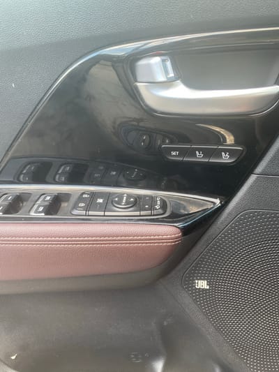 The image shows the interior of a car, specifically the driver's side door panel. The primary objects visible are car controls, including window controls, seat adjustment buttons, and a mirror adjustment control. The seat adjustment buttons are arranged vertically, with a "SET" button at the bottom and two other buttons above it, likely for saving and recalling seat positions. Next to these are the window controls, featuring individual buttons for each window, with "AUTO" labeled on some. Above the window controls, there's a circular button labeled "MIRROR" and a directional control for adjusting the mirrors.

The scene is indoors, likely within the car. There are no people visible. The lighting suggests it's daytime, with sunlight reflecting off the glossy black trim of the door panel. The car appears to be modern, with a sleek design and brown leather upholstery visible on the armrest. A prominent detail is the "JBL" logo on the textured speaker grille, indicating the car has a JBL sound system. The environment is clean, with no obvious signs of damage or disarray.

The overall impression is a close-up shot of the car's interior controls, highlighting the functionality and design elements. The absence of any specific event or action suggests this is a descriptive photograph of the vehicle's interior features. The location context of Rezina, Moldova, provides a geographical reference for where this image was taken, but there are no visual cues within the image to confirm this location.