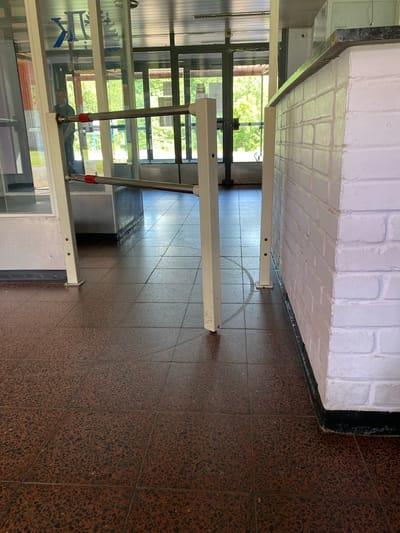 The image depicts an interior hallway with tiled flooring. On the left, there are glass-paneled walls with some blue lettering visible. Two metallic poles with red accents are extending upwards from the floor, supporting a white horizontal bar. In the distance, through the glass panels, trees and sunlight are visible, suggesting it is daytime and the weather is clear. A white brick wall with dark grout is present along the top of the image. The overall scene appears to be a public or commercial space, possibly a hallway in a building or an educational institution. There are no visible people actively engaged in an activity. The lighting suggests natural light entering from the outside. The flooring is a mottled brown and black tile.