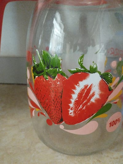 The image is a close-up shot of a glass pitcher with a strawberry design. Two stylized strawberries, one whole and one halved, are depicted in red and green. There are also abstract shapes in pink, yellow, and green surrounding the strawberries. In the bottom right corner, the text "100%" is visible within an oval shape. The pitcher appears to be sitting on a speckled countertop. The background is a light gray surface. The pitcher has a red handle and rim, which are partially visible on the left side of the image. The overall impression is that of a decorative item, likely used for beverages. The context provided regarding "Mady, Kyrgyzstan" does not appear to be directly represented within the visual elements of the image itself.