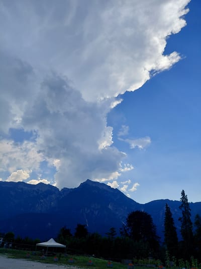 The image captures a mountainous landscape under a dramatic sky, likely in or near Levico Terme, Italy, given the context. The foreground shows a flattened area with a white canopy tent set up, indicating a potential outdoor event or gathering space. Several small, orange-topped objects are arranged in a line near the tent, possibly for decoration or as markers. Lush green vegetation, including trees and grass, forms a dense border behind the open area and extends up the slopes of the mountains.

The mountains themselves dominate the background, their dark, silhouetted forms rising steeply and disappearing into the clouds. The peaks are rugged and textured, with visible crevices and slopes suggesting a significant geological formation. The sky is a striking contrast of deep blue and massive, billowing clouds. A large, white cumulus cloud formation stretches across the upper left and center of the frame, casting shadows and creating a dynamic, textured ceiling to the scene. The right side of the sky is a clear, vibrant blue, with subtle rays of sunlight piercing through the lighter cloud sections.

No people are clearly visible or engaged in any discernible actions within the frame. The environment appears to be a natural setting, potentially a park or recreational area adjacent to the mountains. The overall impression is one of natural grandeur combined with a touch of human presence through the erected tent.