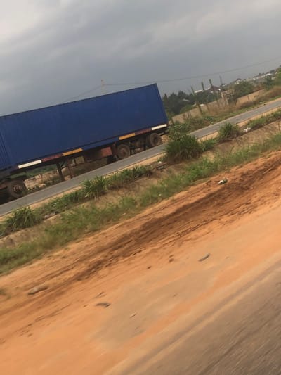 The scene depicts an outdoor environment in Nwofia, Nigeria, under an overcast sky. The foreground comprises reddish-brown earth, slanting from the bottom-right towards the upper-left, marked by subtle undulations and darker streaks. Parallel to this, a narrow, dark grey asphalt road runs diagonally across the mid-ground.
Adjacent to the road, on both sides, are strips of green, dense, low-lying vegetation. A large, dark blue shipping container, affixed to a brown trailer chassis with multiple axles and dark tires, occupies the shoulder or a parallel unpaved section of the road in the middle-left of the image. The trailer features visible yellow and white reflective striping along its lower edge. No individuals are present.
In the background, beyond the road and vegetation, are sparse trees and scattered structures, including what appear to be partially constructed or residential buildings, defined by light-colored walls or concrete elements. Some rudimentary fencing or wall structures are visible. A faint, thin pole or wire extends vertically against the cloudy sky in the far left background. Litter, including white plastic fragments, is visible on the reddish earth near the road.