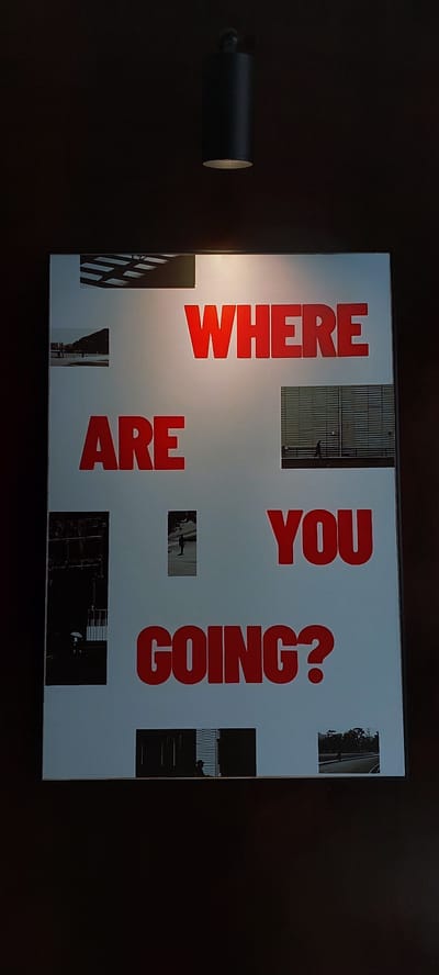 A framed artwork is displayed on a dark wall. The artwork features large, red, sans-serif text on a light-colored background, arranged in three lines. The text reads "WHERE ARE YOU GOING?". Interspersed among the text are several small, black and white photographs. One photo shows architectural elements of a building. Another depicts a single figure walking in front of a modern building. A third photo shows a person under an umbrella. Other photos show silhouetted figures in doorways and a street scene. A dark, cylindrical light fixture hangs from the ceiling directly above the artwork. The environment appears to be indoors, possibly a gallery or public space. There is no specific indication of the location being Pagedangan, Indonesia.