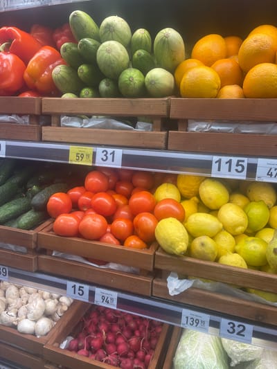The image shows a grocery store produce section, specifically at the Silpo supermarket in Гожули, Ukraine. The focus is on several wooden crates filled with fresh fruits and vegetables.
In the top crates, there are bright red bell peppers on the left, followed by green zucchini and round, light green squashes. To the right of the squashes are various citrus fruits, primarily oranges and some smaller tangerines.
The middle crates contain an assortment of vegetables. On the left, there are dark green cucumbers. Next to them are red tomatoes. To the right of the tomatoes are bright yellow lemons.
The bottom crates display mushrooms on the left, followed by red radishes in the center. On the far right, a head of iceberg lettuce is visible.
Price tags are visible above each section of produce, indicating prices in Ukrainian Hryvnia (UAH). For example, a tag above the cucumbers and tomatoes reads "59 90" and "31", while a tag above the lemons reads "115". Another tag above the mushrooms and radishes shows "15" and "94 90", and the tag above the lettuce shows "139 90" and "32". The lighting appears to be typical supermarket fluorescent lighting. There are no people visible in the image.