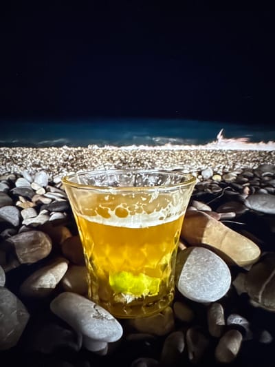 A glass of beer sits on a rocky surface, with a dark body of water and the faint glow of lights in the background. The scene is lit from above, casting shadows on the pebbles surrounding the glass. The beer is golden yellow with a frothy head and a wedge of lime at the bottom of the glass. The background water appears to be choppy, suggesting a windy or rough sea. The overall impression is one of a serene evening at the beach, with the drink adding a touch of relaxation to the atmosphere. There is no identifiable location in the city of Porrentruy, Switzerland.