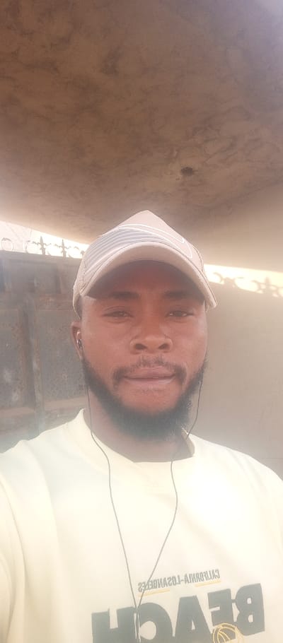 A close-up portrait of an adult male in Tunga Mekidi, Nigeria, is depicted. The individual has dark skin, a full beard, and is wearing a beige baseball cap with light-colored stripes on the front panel. He is dressed in a light-colored long-sleeved shirt, on which the reversed text "BEACH" and "CALIFORNIA-LOS ANGELES" is partially visible. Black wired in-ear headphones are in his ears, with the wire extending downwards. The man looks directly into the camera with a neutral to slight smile. The background features a dark, possibly metallic or weathered, gate structure to the left of the subject, and a plain, light-colored wall or surface to his right. Above the subject is a concrete or similarly structured overhang. The scene is illuminated by ambient daylight.
