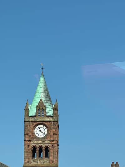 The image shows a close-up view of the historic Guildhall clock tower in the city of Derry/Londonderry, United Kingdom. The clock tower is constructed of red sandstone and features a green, verdigris-covered conical roof topped with a weather vane shaped like a ship. A large clock face with Roman numerals is visible on the tower, with the time appearing to be approximately 10:55. The tower has arched windows and decorative stonework. The sky above is a clear, bright blue. There are no people or animals visible in the frame. The bottom of the image shows a sliver of what appears to be another building with chimneys. The overall scene is one of an architectural landmark against a clear sky.