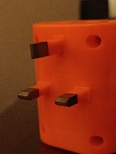 The image shows a close-up of an orange power plug with three prongs. The prongs are rectangular and appear to be made of metal with a black coating. The plug is oriented vertically, with the prongs pointing towards the left. The background is a textured, brown surface. There is a circular indentation on the right side of the plug. The scene is a close-up, forensic, and matter-of-fact depiction of the object. There are no people or actions visible, and the environment is limited to the immediate surroundings of the plug. The location in the city of Keer, Pakistan, is not discernible from the image itself.