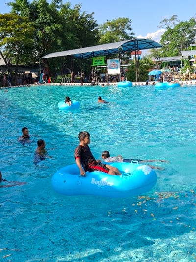 The image shows a swimming pool in a water park located at coordinates -7.592 latitude, 110.649 longitude in Kecamatan Tulung, Indonesia. There are people swimming and floating on blue inflatable tubes in the pool. A young man is sitting on a blue tube with the word "Boulder" written on it. The man is wearing a black T-shirt and red shorts. Other people are swimming in the water, including a man in a red shirt. The scene is sunny and appears to be during the day. The water is clear and blue, and the pool is surrounded by trees and a white metal roof structure. The scene appears to be a water park with a designated area for swimming and floating.