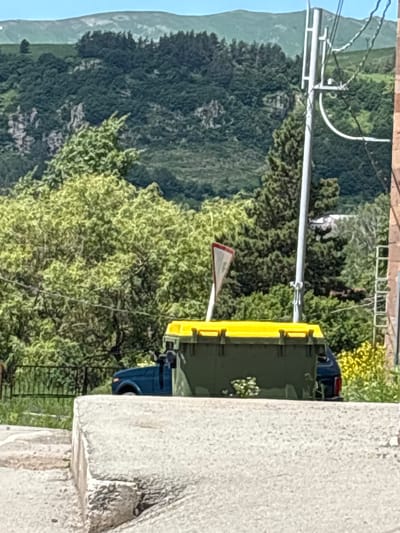 In Jermuk, Armenia, a dark blue pickup truck is parked behind a large, green dumpster with a yellow lid. The dumpster is situated on a paved surface, possibly a street or alleyway, partially obscured by a concrete barrier in the foreground. Behind the truck and dumpster, a dense forest covers rolling hills, leading up to a distant mountain range under a clear blue sky. A power pole with wires runs vertically on the right side of the frame, and a red and white triangular yield sign is visible amongst the trees. The overall scene suggests a quiet, possibly rural or semi-urban environment within the city.
