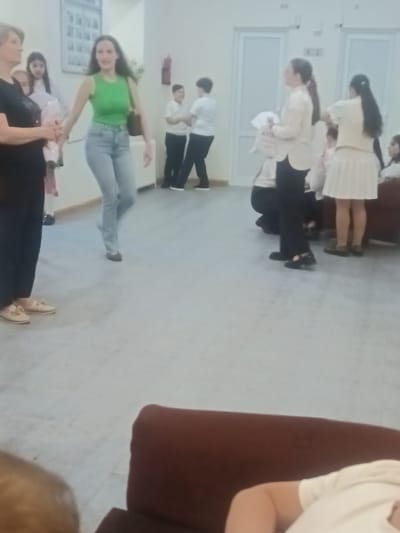 In Nor Hachin, Armenia, a group of individuals, predominantly young women and girls, are gathered in an indoor space resembling a community hall or school corridor. The floor is a light gray tiled surface. On the left, a woman in a black shirt and dark pants, with blonde hair, stands holding hands with a young girl in a light-colored dress. Next to them, a woman with long dark hair wears a vibrant green sleeveless top and light blue jeans, also holding hands with the girl.

Further back, several individuals in white shirts and dark pants or skirts are visible. Two boys in white shirts and dark pants stand facing each other, possibly interacting. To the right, a woman in a light-colored collared shirt and dark pants holds a white item, possibly a piece of fabric, while another woman in a white top and a light-colored pleated skirt stands beside her. The background features cream-colored walls and light-colored doors. The overall atmosphere appears to be a casual gathering or event.