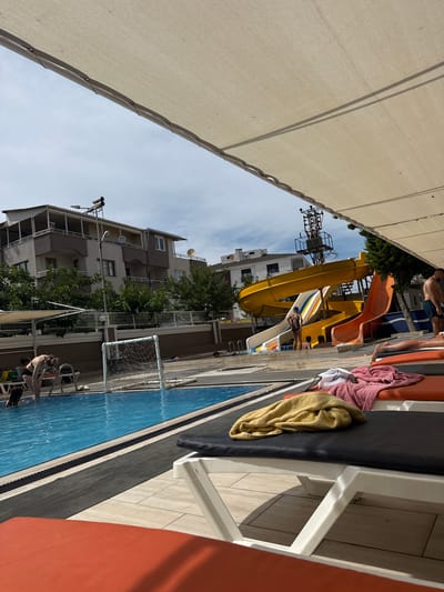 The image depicts a sunny day at an outdoor water park in Ayvalık, Turkey. In the foreground, several lounge chairs are set up, some with towels draped over them. A pool with blue water is visible, with a white water polo goal net in the middle. In the background, a multi-level building with balconies and trees surrounds the water park. A colorful water slide complex with yellow and orange tubes is a prominent feature, with a few people visible on and around the slides. The overall scene suggests a recreational area designed for enjoyment and relaxation.