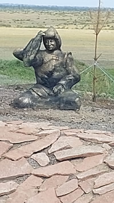 A bronze statue of a seated figure in traditional attire is positioned in an outdoor setting with a dry, grassy landscape in the background. The figure is wearing a helmet and appears to be in a contemplative pose, with one hand raised to its forehead. The statue is placed on a mound of earth, surrounded by green grass, and in the foreground, there is a paved area made of irregular, reddish-brown stones. A thin tree with minimal foliage stands to the right of the statue. The overall scene suggests a historical or cultural monument in a somewhat arid environment. The location is identified as Zhartas, Kazakhstan.