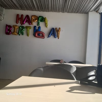 A photograph captures a wall decorated with colorful letter balloons that spell out "HAPPY BIRTHDAY". The balloons are arranged vertically in two columns. The left column spells "HAPPY" with "H" in gold, "A" in magenta, "P" in red, "P" in gold, and "Y" in green. The right column spells "BIRTHDAY" with "B" in green, "I" in blue, "R" in red, "T" in yellow, "H" in blue, "D" in red, "A" in yellow, and "Y" in magenta. The background is a plain white wall with some faint marks and scratches.

To the left of the balloons, a grey, pleated curtain is visible. To the right of the balloons, there are several large, dark grey chairs, possibly office chairs, stacked on top of each other. A small white tag with the number "33" printed in red is attached to one of the chairs. The lighting suggests it is daytime, with strong shadows cast from the chairs onto a light-colored surface to the right of the image. The text "Galaxy A14" is visible on this surface, indicating the device used to take the photo. The scene appears to be indoors, perhaps an office or event space. There are no people visible in the image. The overall mood is festive due to the balloons.