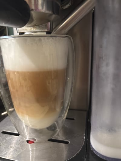 A close-up, eye-level shot captures a glass of a layered coffee drink being prepared by an espresso machine. The drink features distinct layers of white foam at the top, followed by a tan coffee layer, and a milky white base. The glass is transparent and appears to have a double-walled design. The espresso machine's metallic components, including a black handle and a chrome spout, are visible at the top of the frame. To the right, a clear plastic container, possibly for milk or water, is partially visible, showing a white liquid inside. The machine rests on a metallic surface with visible textured details. A small red button or indicator is positioned below the glass. The overall environment appears to be a kitchen or coffee-making area. There is no indication of the location being in Zubov, Russia.