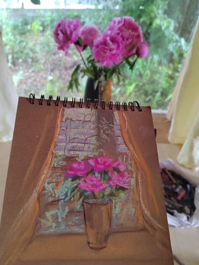 The image shows a close-up view of a sketchbook with a pastel drawing of a vase of pink flowers. The drawing depicts the flowers in a brown vase against a background that appears to be a brick wall and curtains. The sketchbook itself has a spiral binding and is placed on a surface, possibly a table. In the blurred background, out of focus, a bouquet of real pink flowers is visible in a vase, with greenery and trees seen through a window. A bag filled with art supplies, likely pastels, is also visible to the right of the sketchbook. The setting suggests an indoor art studio or a room with a view of nature. There is no identifiable location in the city of Бярэзінскі сельскі Савет, Belarus.