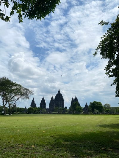 The image shows the Prambanan Temple complex, a UNESCO World Heritage Site located in Kecamatan Prambanan, Indonesia. It's a sunny day with a blue sky and white puffy clouds. The temple is silhouetted against the bright sky, with the tallest spire in the center. The foreground consists of lush green grass that stretches out toward the temple. There are a few trees on the left side of the image, and a small cluster of trees on the right. A bird is flying across the sky in the center of the image. The scene is peaceful and serene.