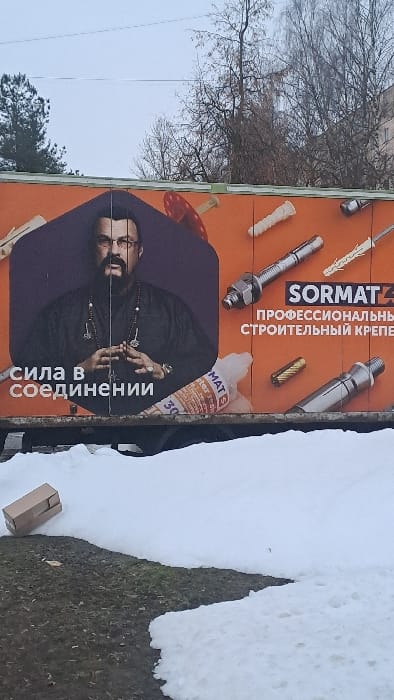 The scene, presented as being in Bel'kovo, Russia, depicts an outdoor environment under an overcast sky. In the foreground, patches of melting snow cover areas of dark, exposed ground, which appears to be dormant grass or dirt. A small, light-brown cardboard box lies horizontally on its side, resting on the ground near a snow patch on the lower left.
Dominating the midground is a large, rectangular advertisement panel, likely mounted on a temporary structure or trailer, positioned above the snow. The advertisement features an orange background. On the left half, a portrait of a man, identifiable as Steven Seagal, is prominently displayed. He wears glasses, a mustache, a goatee, a dark garment resembling a tunic or shirt, and multiple necklaces adorned with pendants. His hands are clasped together at chest level. Below his image, white Cyrillic text reads "СИЛА В СОЕДИНЕНИИ" (Strength in Connection). The right half of the advertisement displays various construction fasteners, including expansion anchors, bolts, and screws. The brand "SORMAT" is visible in white lettering, alongside Cyrillic text "ПРОФЕССИОНАЛЬНЫЙ СТРОИТЕЛЬНЫЙ КРЕПЕЖ" (Professional Construction Fasteners). A partially obscured tube, consistent with a sealant or adhesive product, is also depicted.
The background shows a light, overcast sky. Bare deciduous trees stand on the right, while a dark green coniferous tree is visible on the far left. A light-colored, multi-story building is partially visible through the bare branches on the right side of the image.