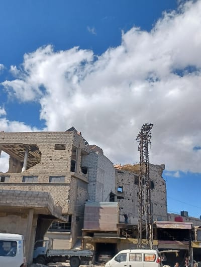 The image depicts an urban street scene in Al-Qadam Municipality, Syria, under a blue sky with scattered white cumulus clouds. The central focus is a cluster of multi-story buildings exhibiting extensive structural damage, characterized by collapsed sections, exposed interiors, missing windows, and numerous impact holes consistent with bullet or shrapnel strikes across their concrete block facades. A lone individual is visible standing within an opening on the second floor of the central damaged building.

A tall, rusty metal lattice transmission tower stands upright in the mid-ground, positioned between the damaged structures and the street level. On the street, a white van is partially visible on the far left, with a driver discernible inside. A white pickup truck is parked left of center. To the right, a white passenger van is parked, and a dark vehicle is visible inside a ground-level garage entrance. Two damaged commercial storefronts are present on the right, one featuring Arabic script above its entrance and a red cylindrical tank on its roof. Another individual is partially visible near the white passenger van on the right, appearing to be sitting or crouching.