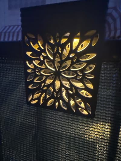 A dark, decorative solar lantern hangs on a dark green mesh fence. The lantern has a cut-out floral pattern that allows a warm yellow light to emanate from within. The background shows a blurry, striped awning in shades of blue and white. The scene appears to be outdoors, likely at dusk or night, given the illumination from the lantern. The location is identified as Santa Pola, Spain.