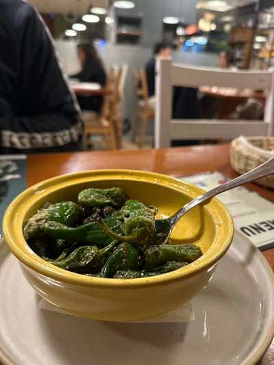 A yellow bowl filled with sautéed green peppers sits on a white plate. A silver fork rests in the bowl amongst the peppers. The bowl and plate are on a table. In the background are chairs and people in a dimly lit restaurant. Part of a menu can be seen behind the bowl. A person wearing a black jacket with white stripes is seated at the table.