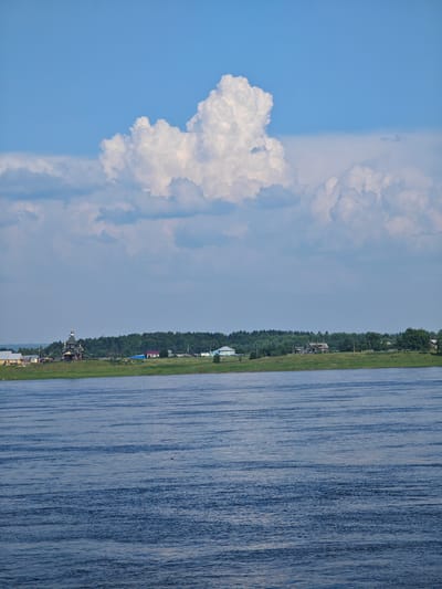 A wide, blue river flows in the foreground, its surface rippled by the wind. Beyond the river, a lush green grassy bank stretches across the scene, dotted with trees. Further inland, a cluster of buildings forms a small settlement. These include several houses, some with red roofs, and a prominent, dark wooden church with a golden dome, situated on a slight elevation. A dense forest forms the backdrop to the settlement. The sky above is a clear blue, marked by a large, puffy white cloud formation. The image appears to capture a serene rural landscape in Verkhnepashino, Russia.