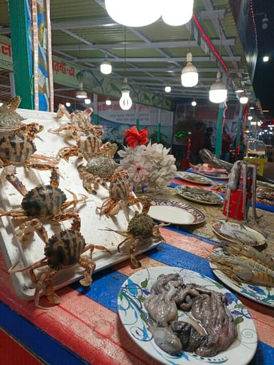 A seafood display features a variety of fresh catches. Crabs with distinctive striped patterns are arranged on a white stand. Nearby, a plate holds uncooked octopus, and another plate displays prawns. Additional fish and seafood items are scattered on the table. A white floral arrangement with a red bow is positioned between the crabs and the other seafood, adding a decorative touch. The display is set beneath bright overhead lights.