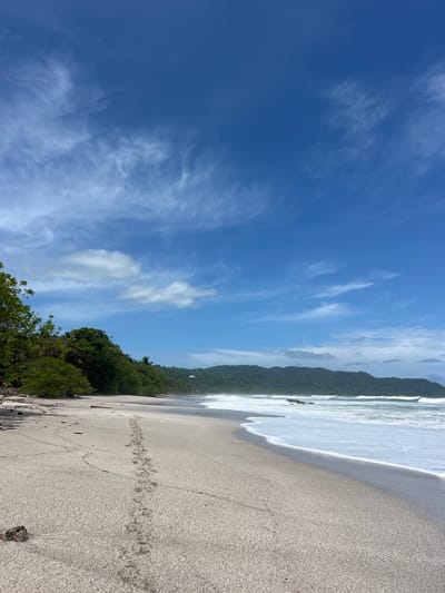 The image shows a beach with white sand and foamy waves breaking on the shore. The beach is mostly empty, with only a set of footprints in the sand extending toward the water. There is a line of dense green vegetation growing along the shoreline, followed by a range of forested hills in the distance. The sky is a bright blue with white wispy clouds. The location of the image is Playa Naranjo in Santa Teresa, Costa Rica.