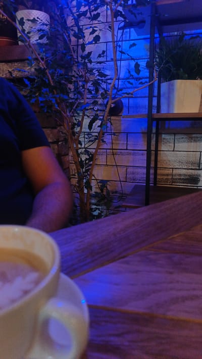 The image depicts an indoor scene, likely a cafe, with a person seated at a table. The dominant lighting is a deep blue, creating a moody and atmospheric effect. In the foreground, a portion of a light-colored mug filled with a light-colored beverage is visible, resting on a saucer and placed on a wooden table. The table has a visible grain and appears to be made of planks.
To the left, the arm and shoulder of a person wearing a dark blue or black t-shirt are visible. The person's arm is positioned at an angle, suggesting they are seated.
In the background, a thin, artificial tree with sparse green leaves is prominent. Behind the tree, a brick wall is visible, partially obscured by the foliage. The bricks are painted in shades of white and grey, with dark lines indicating the mortar. The blue lighting casts shadows and highlights on the wall, emphasizing its texture.
To the right of the tree, a metal shelving unit with wooden shelves is present. On the shelves are potted plants. One pot, light in color, holds a plant with dense green foliage. Another plant, with thin, grass-like leaves, is in a white pot. The shelves and their contents are illuminated by the blue light.
The overall scene suggests a relaxed or intimate setting. The blue lighting could indicate evening or night time. There are no visible text elements in the image. The context provided places this scene in Langepas, Russia.