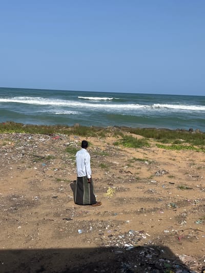In Moratuwa, Sri Lanka, a man wearing a white shirt and a dark sarong stands on a sandy beach littered with trash. The ocean waves crash in the background under a clear blue sky.