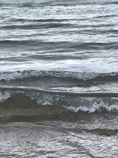 The image captures a close-up view of the sea with multiple waves. The water is a muted gray color, and the waves are rolling in with crests of white foam. The lighting is overcast, giving the scene a somber and somewhat monochromatic feel. The composition is abstract, focusing solely on the movement and texture of the water. There are no people, animals, or man-made objects visible in the frame. The location is the North Sea coast in Bergen, Netherlands.