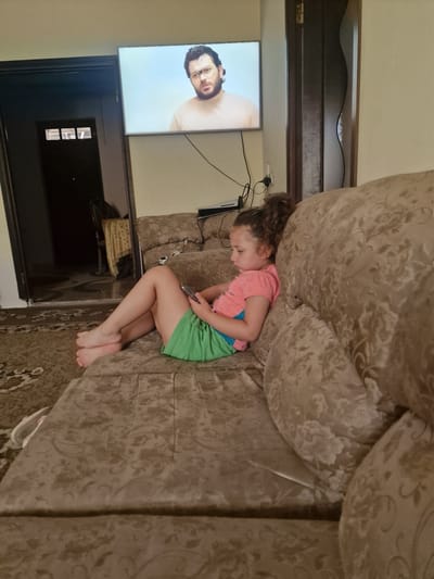 In the city of Nor Kharberd, Armenia, a young girl is seen sitting on a patterned couch, engrossed in her mobile phone. She is wearing a pink t-shirt and green shorts. The couch is positioned in a living room, with a television screen displaying an image of a man wearing glasses in the background. A doorway is visible to the left of the television. The room is lit, creating a casual, indoor atmosphere. The rug on the floor has a complex pattern.