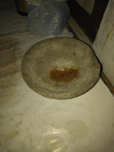 The scene, located in a building within Tarqumiyah, Palestinian Territory, features a circular, shallow, grey stone vessel, approximately 20-25 cm in diameter, resting on a light-colored floor. The stone's surface is coarse, and its interior contains a central, reddish-brown, circular stain, consistent with dried residue. The surrounding floor exhibits multiple irregular yellowish-brown stains. In the upper left background, a metallic cylinder and a crumpled, translucent grey plastic bag are partially visible. Dark, indistinct shapes occupy the upper right background. No individuals are present, and no actions are depicted.