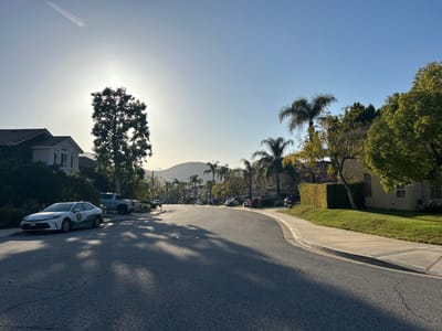 The image shows a residential street in Oak Park, United States of America. The street curves gently to the right, with sidewalks on both sides. Houses line the street, some with manicured lawns and hedges. A variety of trees, including palm trees, are scattered along the street. Several parked cars are visible. A mountain is discernible in the distant background. The sun appears to be low, casting long shadows across the road. The sky is a clear, light blue. There is a contrail in the sky to the left. The asphalt road shows signs of wear and tear.
