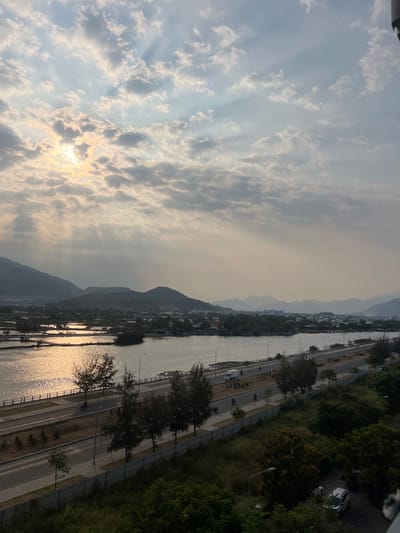 The image shows a landscape with a river, mountains, and city elements. The sky is filled with clouds, and the sun shines through them. The river reflects sunlight. A road runs along the river's edge, with trees and some buildings. The mountains are visible in the background. The location is in or near Nam Natural Heritage Area Trang, Vietnam.