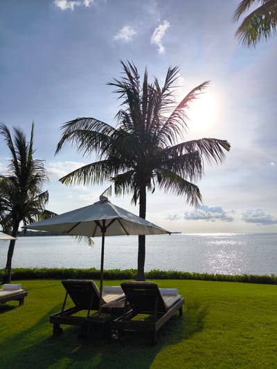The image depicts a tranquil beach scene in Sigar Penjalin, Indonesia. A well-manicured green lawn slopes gently towards a body of water, likely the ocean, shimmering under a bright sun. Two dark-wood lounge chairs with cushions sit beneath a large white umbrella, suggesting a relaxing spot. A tall palm tree, partially obscuring the sun, casts shadows on the lawn. Another palm tree is visible on the left, partially out of frame. The sky is pale blue with scattered white clouds. The sun's glare creates a sparkling effect on the water's surface. A hint of land or an island can be seen in the distant background.
