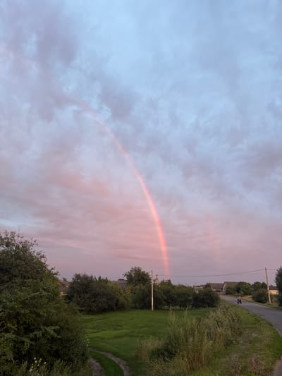 The image depicts a rural landscape in Istobnoye, Russia, under a twilight sky. A prominent double rainbow arches across the sky, with soft pink and blue hues dominating the clouds. In the foreground, lush green grass and trees create a natural border. A dirt path curves through the grass, leading towards a paved road. On the road, a person is riding a motorcycle, heading away from the viewer. Several houses with tiled roofs are visible in the middle ground, interspersed with dense foliage. Power lines and utility poles are present, indicating a connection to infrastructure. The overall atmosphere is serene, capturing the beauty of nature at dusk.