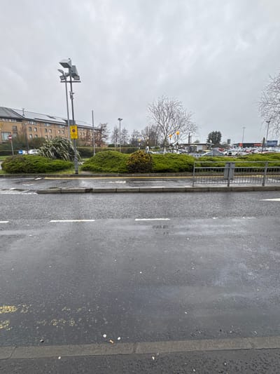The image depicts an overcast outdoor scene in Crumlin, United Kingdom, characterized by a wet asphalt roadway and surrounding commercial infrastructure. The foreground features a dark grey asphalt road, visibly damp from rain, with two sets of broken white lane markings and faded yellow double-line markings along the curb. Scattered litter, including several cigarette butts, is present on the asphalt and the concrete curb. A low, dark grey metal pedestrian barrier extends from the right edge into the midground.
In the midground, a landscaped central reservation or roundabout is covered in dense green shrubbery, with several deciduous trees showing bare branches. A tall, dark grey utility pole with multiple light fixtures and an attached surveillance camera or sensor array stands to the left of the center. Attached to this pole are a yellow circular sign indicating "No Waiting" and a blue circular sign with a white arrow pointing left. To the left, a multi-story building with a light brown or beige facade and numerous windows is visible, identified in part as a "Premier Inn" by partial signage; its roof features a section of solar panels.
The background shows a large car park filled with various vehicles. A prominent "Tesco" supermarket sign is visible above the car park on the right side. The sky is uniformly grey and overcast. No individuals are visible, and no specific actions or interactions are depicted within the scene.