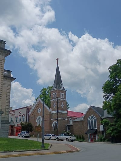 The image shows a street scene in Paris, United States of America, featuring a church and adjacent buildings under a bright, cloudy sky. On the left, a portion of a large, stone building with classical architectural elements is visible. To its right is a brick church with a tall steeple topped by a red cross. The church has arched windows and decorative brickwork. In front of the church, there is a paved road with a yellow curb and a grassy area. A white police SUV with "FIRE DEPT." marked on its side is parked on the right side of the road, facing away from the camera. Another white car is parked further down the street. The street appears to be in a town or city center. The overall impression is of a serene day in a historic urban environment.